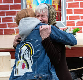 Elderly woman embraces Virgin Mary, against a painted brick background.